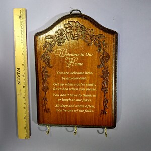 May include: A wooden key holder with a carved floral design and the text "Welcome to Our Home". The text below reads "You are welcome here, be at your ease. Get up when you're ready, Go to bed when you please. You don't have to thank us or laugh at our jokes. Sit deep and come often, You're one of the folks."