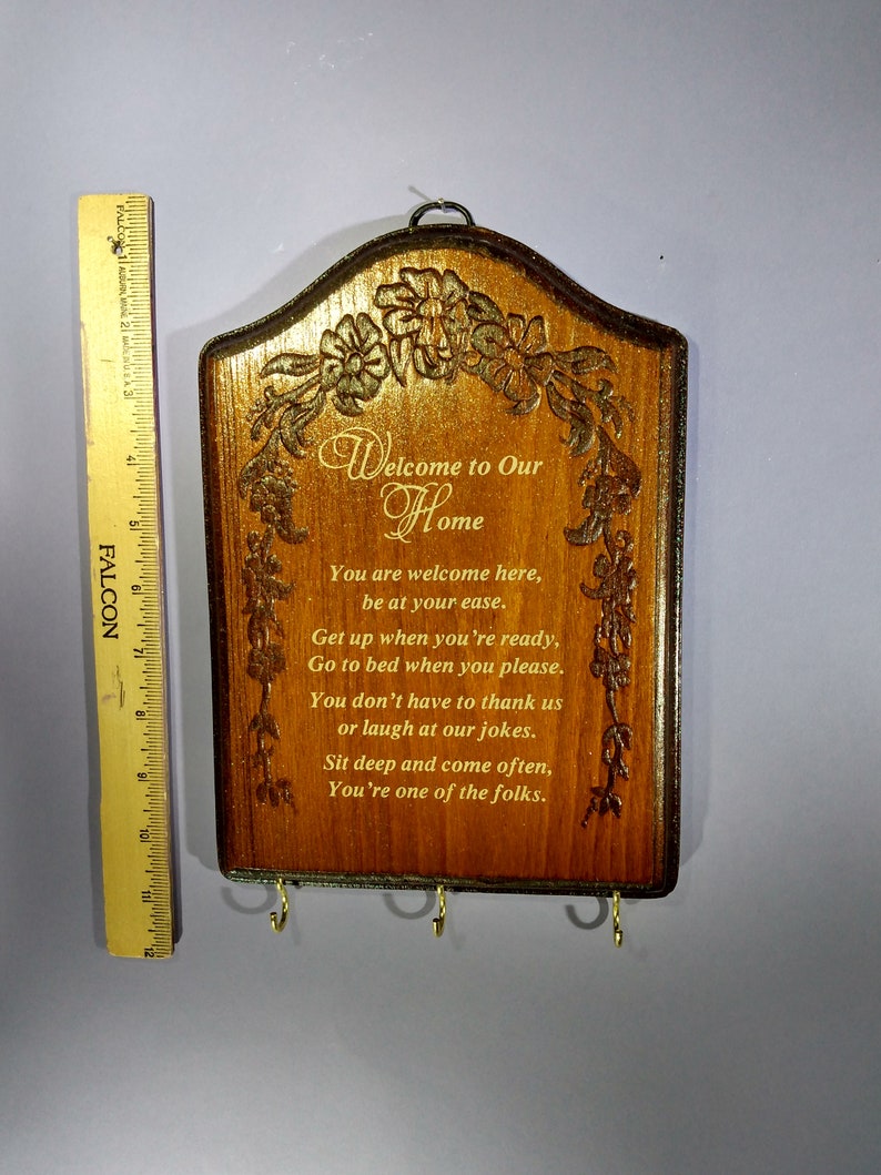 May include: A wooden key holder with a carved floral design and the text "Welcome to Our Home". The text below reads "You are welcome here, be at your ease. Get up when you're ready, Go to bed when you please. You don't have to thank us or laugh at our jokes. Sit deep and come often, You're one of the folks."