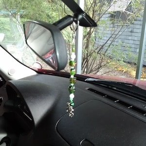May include: A green and white beaded car charm hangs from the rearview mirror of a car. The charm features a small silver charm at the bottom.