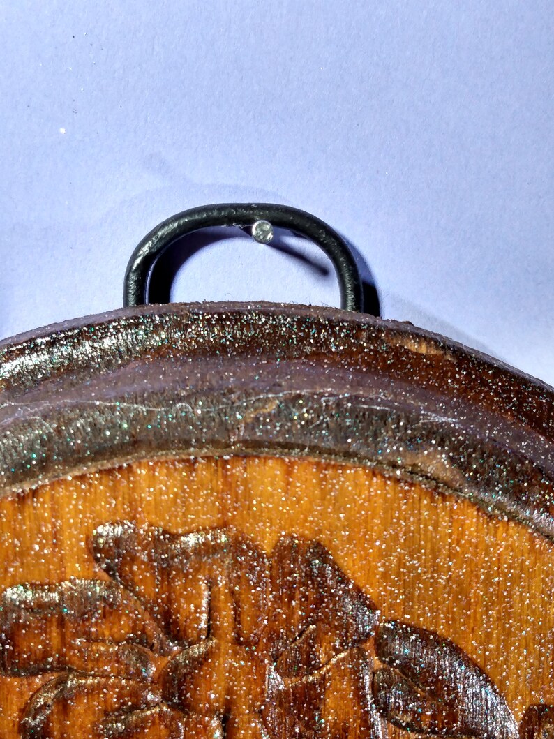 May include: A wooden round ornament with a glittery brown finish and a carved floral design. The ornament has a black metal hook on the top.
