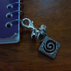 May include: A silver charm with a spiral design and a silver clip. The charm is attached to a purple notebook.