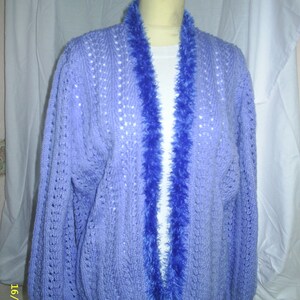 May include: A lilac knitted cardigan with a deep blue, fluffy trim along the open front. The cardigan features a textured knit design and long sleeves. The garment is displayed on a mannequin.