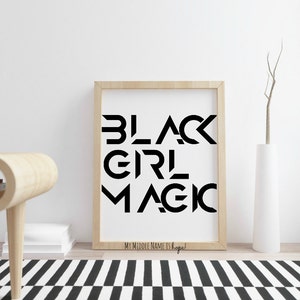 Pode incluir: Uma impress&atilde;o emoldurada em preto e branco com o texto "BLACK GIRL MAGIC" em uma fonte geom&eacute;trica em negrito. O texto est&aacute; centralizado em um fundo branco. A moldura &eacute; de cor madeira clara. A impress&atilde;o est&aacute; sobre uma superf&iacute;cie branca com um tapete listrado preto e branco em primeiro plano.