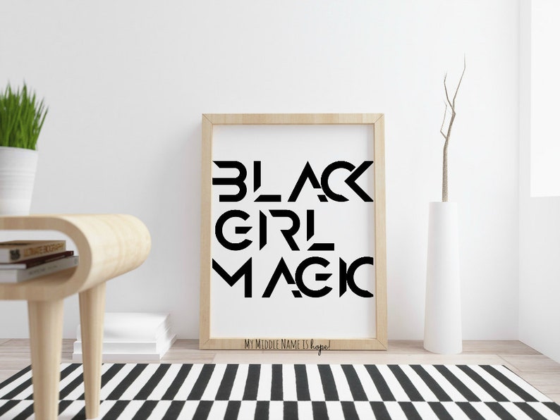 Pode incluir: Uma impress&atilde;o emoldurada em preto e branco com o texto "BLACK GIRL MAGIC" em uma fonte geom&eacute;trica em negrito. O texto est&aacute; centralizado em um fundo branco. A moldura &eacute; de cor madeira clara. A impress&atilde;o est&aacute; sobre uma superf&iacute;cie branca com um tapete listrado preto e branco em primeiro plano.