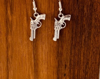 May include: A pair of silver dangle earrings featuring a detailed gun design. The earrings are hanging from a wooden surface.