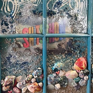May include: A teal window frame containing a collage with the text SHITSHOW in 3D letters. The artwork features silver and blue glitter, various painted stones, and small decorative elements including a stone with the word LIVE and one with PEACE.