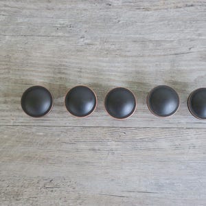 May include: Five black cabinet knobs with a copper-colored trim are arranged in a row against a light gray wooden surface. The knobs have a rounded, dome-like shape, offering a modern aesthetic for furniture.