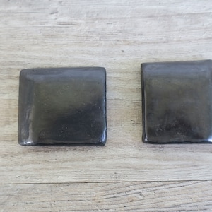 May include: Two square, dark gray ceramic coasters with rounded edges are displayed on a light wood surface. The coasters have a smooth, glossy finish and appear to be handmade. They are designed to protect surfaces from heat and moisture.
