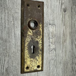 May include: An antique rectangular door plate with a keyhole and a circular opening. The plate has a tarnished brass finish with signs of wear and age, including areas of discoloration and rust. Four screw holes are visible.
