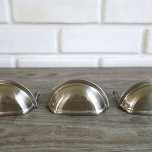 May include: Three brushed nickel-finished drawer pulls in a half-moon shape. Each pull has a small screw for mounting. The pulls are arranged in a row on a gray wooden surface, with a white brick wall in the background.