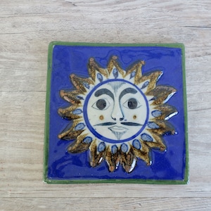 May include: A blue ceramic tile with a hand-painted sun design. The sun has a white face with black eyes, a black mustache, and a black mouth. The sun is surrounded by a gold and brown design.