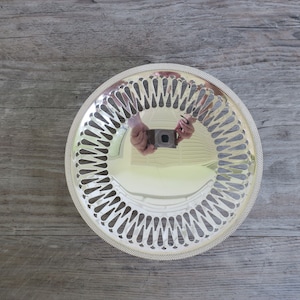 May include: A silver-colored, round decorative tray with a pierced, scalloped edge. The tray has a reflective surface and a detailed, openwork design around the rim. The tray is placed on a wooden surface.