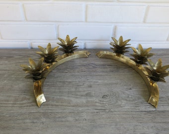 May include: Two brass pineapple-shaped candle holders. Each holder has a curved design with three pineapple accents. The candle holders are made of brass and sit on a wooden surface.
