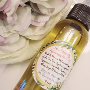 May include: A clear glass bottle with a yellow liquid inside. The bottle has a white label with the text "Low Porosity Ayurvedic Hair Oil" and a list of ingredients including Tea Tree, Jojoba, Grape, Peppermint, Lavender, Rosemary, Clary Sage & Geranium Oil. The label also includes the text "Easy IG: Nourished Life, LLC" and "8 fl oz".