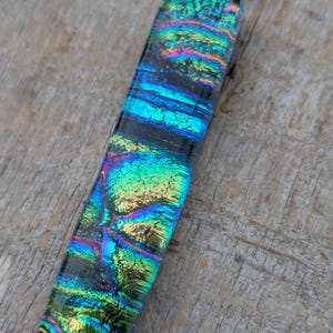 Könnte beinhalten: Ein rechteckiger Anhänger aus dichroitischem Glas mit einem lebendigen, irisierenden Muster in schimmernden Grün-, Blau- und Violetttönen. Das Glas hat eine glatte, glänzende Oberfläche und ist an einer silberfarbenen Metallöse befestigt.