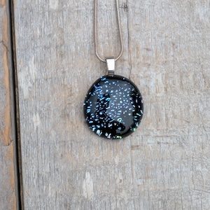 Op de afbeelding: Een ronde, zwarte hangerketting met een zilveren ketting. De hanger heeft een donkere achtergrond met blauwe en iriserende vlekken, wat een sterrenhemel-effect creëert. De ketting wordt getoond op een verweerd houten oppervlak.