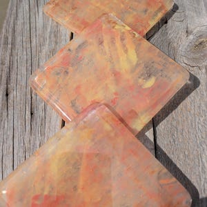 Puede incluir: Tres posavasos cuadrados con un diseño translúcido y veteado en tonos naranja, amarillo y rosa. Los posavasos están apilados uno encima del otro.