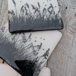 Puede incluir: Posavasos cuadrados de vidrio con un diseño botánico en blanco y negro. Los posavasos presentan una silueta de hierba alta, flores y mariposas. Los bordes de los posavasos son negros y el fondo es blanco.