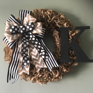 May include: A burlap wreath with a black and white striped ribbon, a black and white polka dot ribbon, and a brown and white chevron ribbon. The wreath is decorated with a black letter "K".