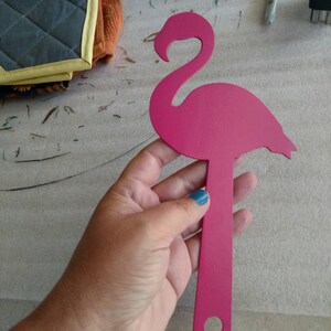 K&ouml;nnte beinhalten: Pinkfarbene Metall-Flamingo-Silhouette mit einem Loch am unteren Ende des St&auml;nders. Der Flamingo wird in einer Hand mit blauem Nagellack gehalten.