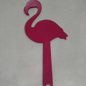 K&ouml;nnte beinhalten: Eine rosafarbene Metall-Flamingo-Silhouette mit einem langen, d&uuml;nnen Stiel. Der Flamingo steht auf einer wei&szlig;en Oberfl&auml;che.