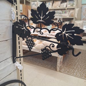 May include: Black metal wall-mounted plant hanger with a grapevine design. The hanger has a hook at the end and a price tag that says "$10.99 Plant Hanger".