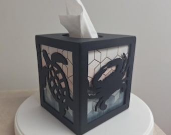 May include: Black tissue box cover with a cutout design of a crab and a turtle. The box is on a white rotating base and has a white tissue peeking out of the top.