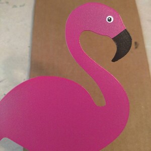 K&ouml;nnte beinhalten: Ein rosafarbener Metall-Flamingo mit schwarzem Schnabel und wei&szlig;em Auge. Der Flamingo steht auf einer braunen Oberfl&auml;che.