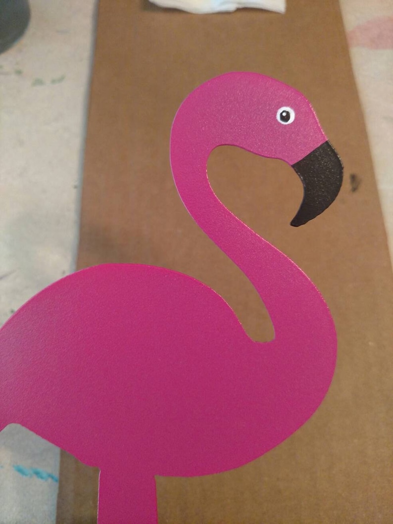 K&ouml;nnte beinhalten: Ein rosafarbener Metall-Flamingo mit schwarzem Schnabel und wei&szlig;em Auge. Der Flamingo steht auf einer braunen Oberfl&auml;che.