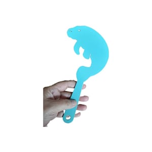 May include: A light blue plastic manatee-shaped kitchen utensil with a handle. The utensil is held in a hand.