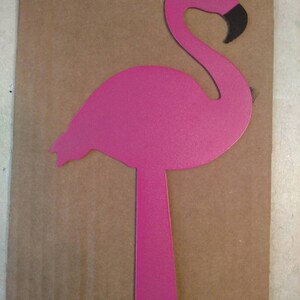 K&ouml;nnte beinhalten: Ein rosafarbener Flamingo-Ausschnitt aus Metall mit schwarzem Schnabel und wei&szlig;em Auge. Der Flamingo steht auf einem Bein mit einem Loch im Boden.