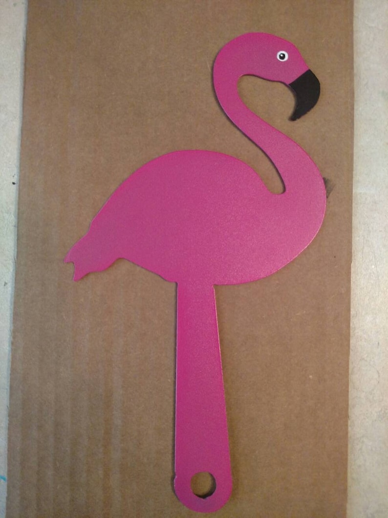 K&ouml;nnte beinhalten: Ein rosafarbener Flamingo-Ausschnitt aus Metall mit schwarzem Schnabel und wei&szlig;em Auge. Der Flamingo steht auf einem Bein mit einem Loch im Boden.