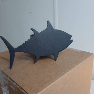 May include: A black metal silhouette of a tuna fish, mounted on a brown cardboard box. The box has a red "Fragile" label.