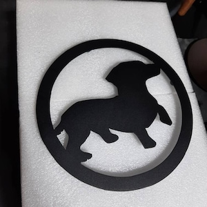 May include: Black metal silhouette of a dachshund dog inside a circle. The silhouette is cut out of the metal and the background is white.