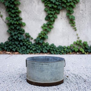 May include: A round, galvanised metal basin with a weathered, grey finish and a dark, rusted band around the base. The container features two small, vertical metal handles attached to the sides, providing a simple and functional design.