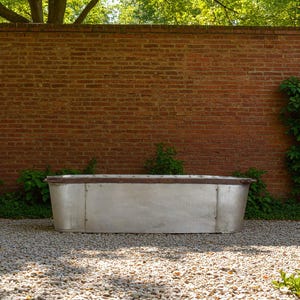 May include: A large, rectangular, galvanised metal trough with a dark brown trim. The trough is set on a bed of small stones in front of a red brick wall. Green foliage is visible on either side of the trough.