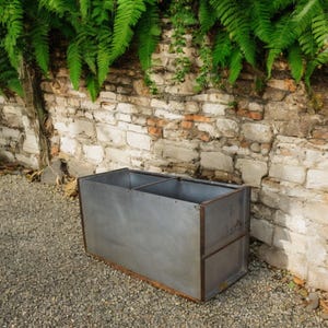 May include: A large, rectangular metal planter with a weathered grey finish. The planter is divided into two sections and sits on a gravel surface in front of a brick wall.