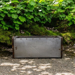 May include: A rectangular metal planter with a weathered appearance. The planter has a silver-coloured metal front and sides, with a darker, possibly rusted, trim. The planter is sitting on a gravel surface, with lush green foliage in the background.