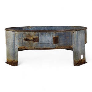 May include: A circular, galvanised metal fire pit with a weathered, rusted finish. The structure features a low-profile design with four integrated, wide legs and a small, rectangular access door on the side secured by a simple metal latch mechanism.