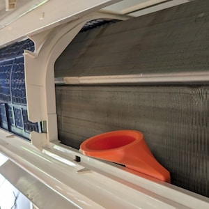 May include: A close-up of an air conditioner unit with a bright orange plastic funnel inserted into the unit. The funnel is used to direct water away from the unit.