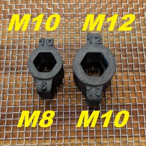 May include: Two black, hexagonal socket adapters are displayed. The larger adapter is labeled "M12" and "12", while the smaller one is labeled "M10" and "10". The text "M8" and "M10" are also present. The adapters are on a metal mesh surface.