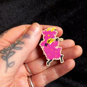 Peut inclure: Une épingle en bois rose avec une chèvre de dessin animé jouant du violon. La chèvre a des yeux et des cornes jaunes et porte une tenue à motifs rose et blanc.