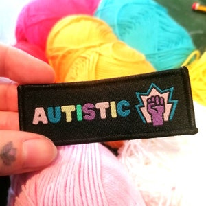 Peut inclure: Écusson en tissu noir avec le mot "AUTISTIC" en couleurs arc-en-ciel et un poing violet levé en l'air avec un contour blanc.