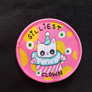 Peut inclure: Un écusson rond brodé rose et jaune avec un chat blanc portant un chapeau de fête et un costume de clown. Le texte "SILLIEST CLOWN" est écrit autour du chat.