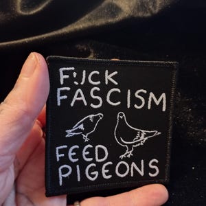 Könnte beinhalten: Schwarzer gestickter Patch mit weißem Text, der "F*CK FASCISM FEED PIGEONS" lautet. Der Patch zeigt zwei weiße Umrisse von Tauben.