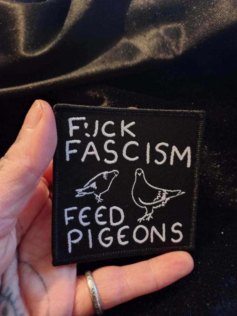 May include: Black embroidered patch with white text that reads "F*CK FASCISM FEED PIGEONS". The patch features two white outlines of pigeons.