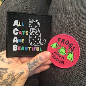 May include: Two embroidered patches. One black patch with a white cat and the text "ALL CATS ARE BEAUTIFUL" in rainbow colors. The other patch is pink with three green frogs and the text "FROGS AGAINST FASCISM".