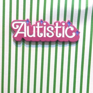 Puede incluir: Pin de esmalte rosa con la palabra "Autistic" en letras blancas y detalles de purpurina azul.
