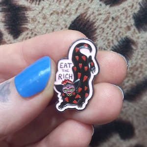 May include: A black and red enamel pin featuring a cartoon monster with the text "Eat the Rich" on a pink background.