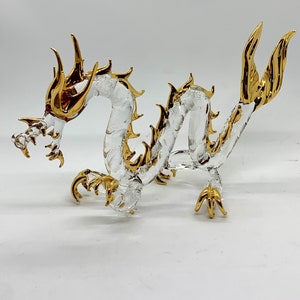 Puede incluir: Una figurita de dragón de vidrio transparente con detalles dorados. El dragón está enroscado con la cola enroscada alrededor de su cuerpo. La cabeza del dragón está levantada y su boca está abierta.